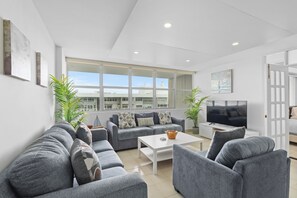 A modern living room with grey sofas, a coffee table, and a TV. There are large windows and potted plants. - Oceanfront Family Retreat 2BR2BA Penthouse Pool & Parking (Carolina)