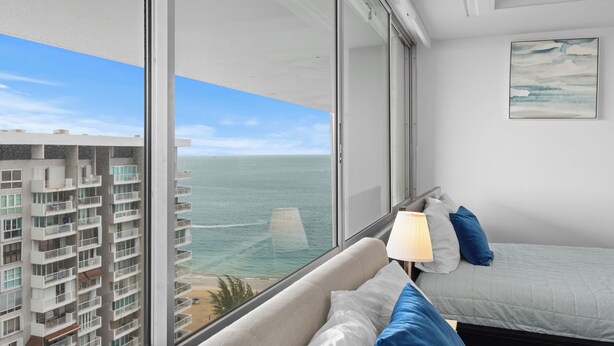 Ein Hotelzimmer mit großem Fenster mit Meerblick, einem Bett mit blau-weißer Bettwäsche und einem Gemälde an der Wand.