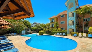 Ein Pool mit Liegestühlen, eine überdachte Sitzgelegenheit und ein mehrgeschossiges Gebäude mit Balkonen.