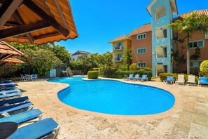 Ein Pool mit Liegestühlen, eine überdachte Sitzgelegenheit und ein mehrgeschossiges Gebäude mit Balkonen.