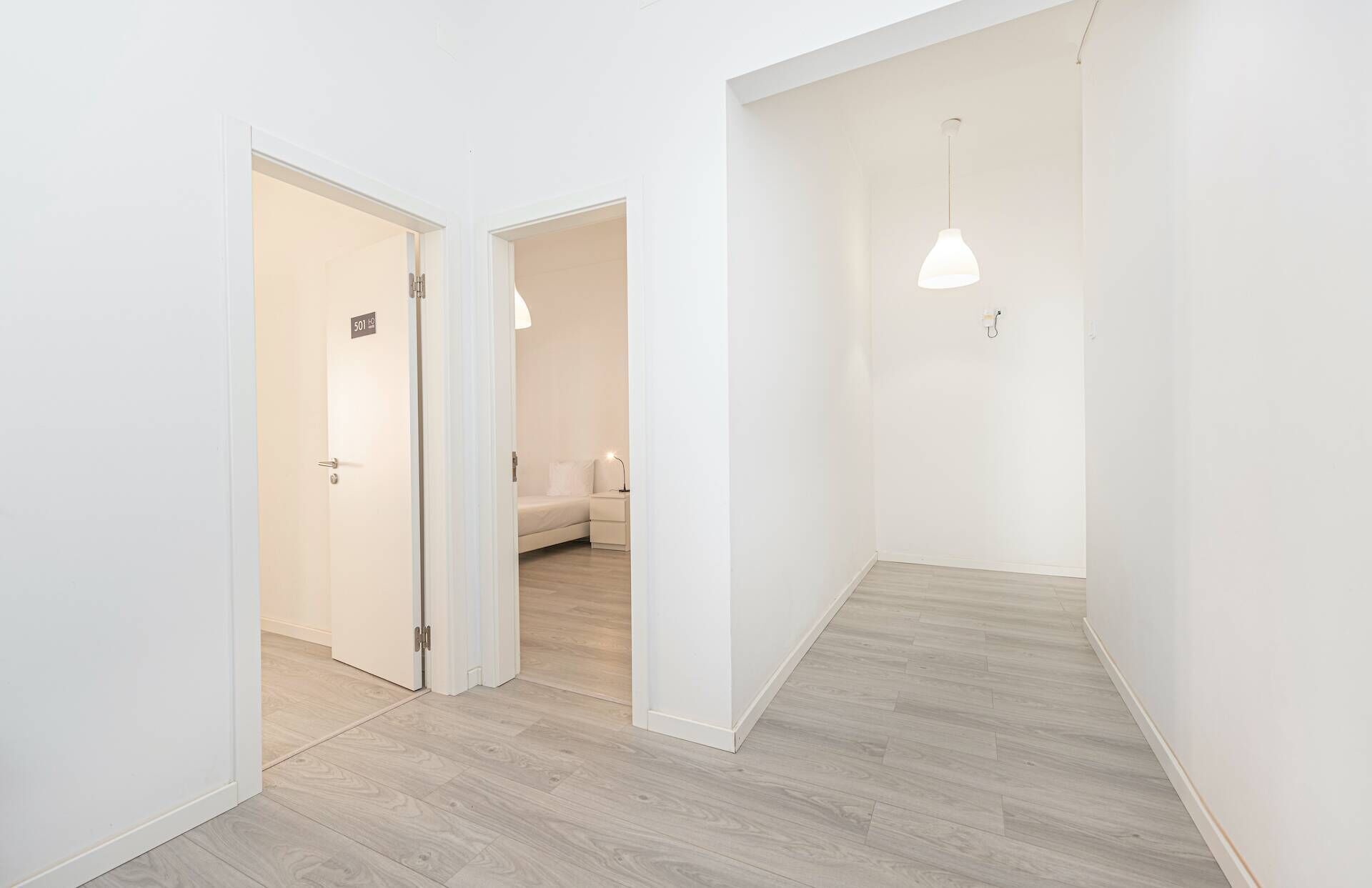 En hall med en dörr märkt med siffran ’501’, som leder in till ett rum med en säng och en lampa.