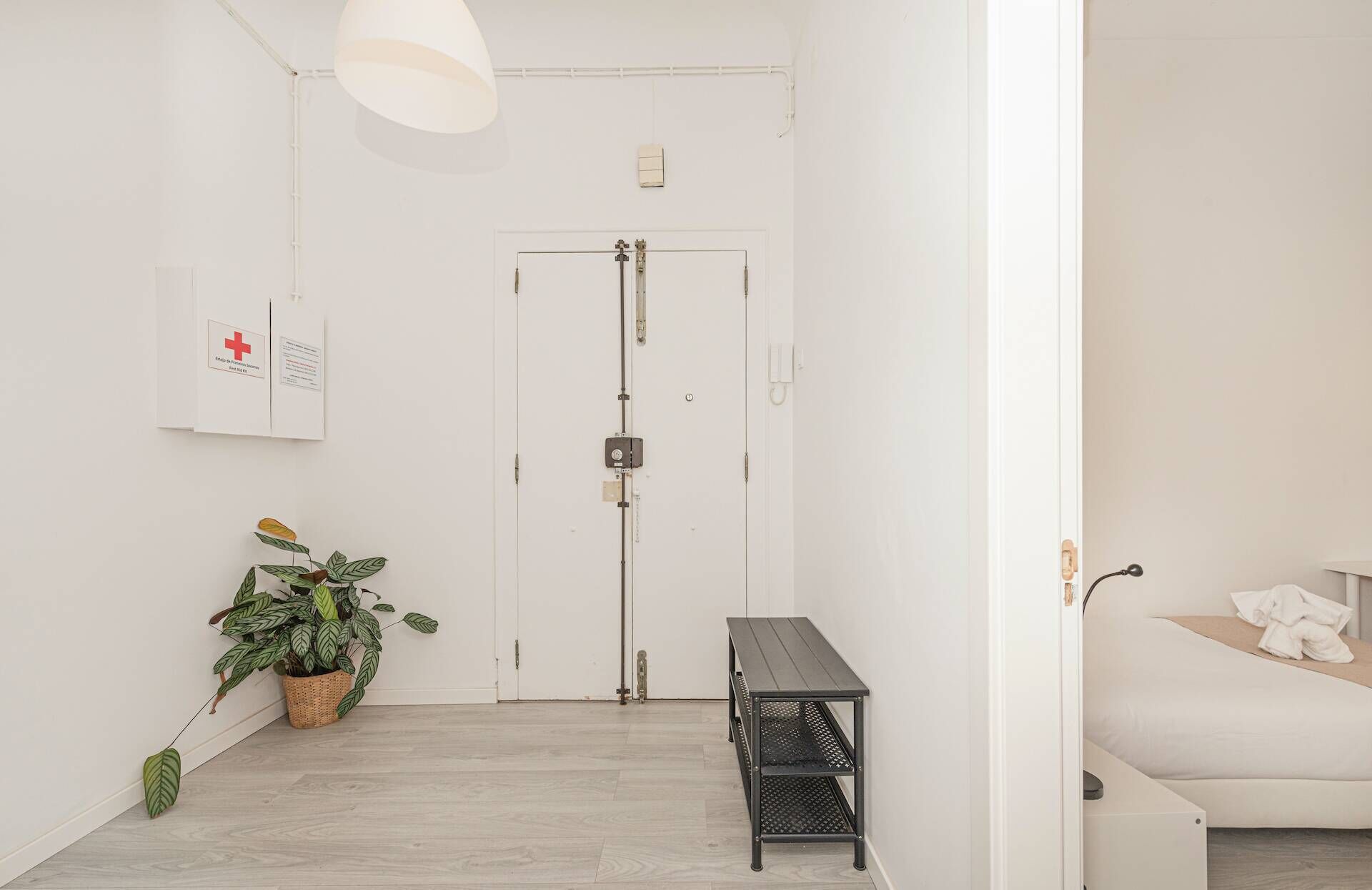 Un pasillo con una puerta blanca, un botiquín de primeros auxilios sujeto a la pared, una planta en maceta y un banco.