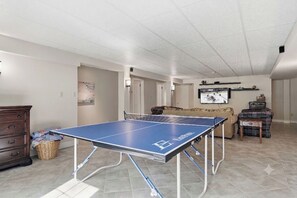 Et bordtennisbord i et rum med flisegulv, en kommode, en kurv og et fjernsyn, der viser en sportsbegivenhed.