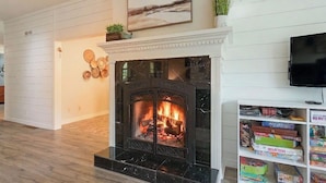 A living room with a fireplace, a television mounted on the wall, and a bookshelf with board games. - WoodPark Cottage 7bedroom Gamesroom (Lambton Shores)