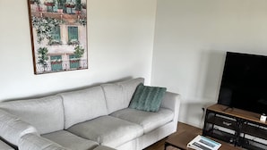 Ein modernes Wohnzimmer mit einem grauen Ecksofa, einem Couchtisch, einem Flachbildfernseher und einem gerahmten Stadtbild an der Wand.