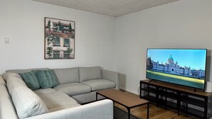 Ein Wohnzimmer mit einem Ecksofa, einem Couchtisch, einem Flachbildfernseher, einem Stadtbild im Bilderrahmen und einem Bücherregal.