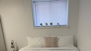 Ein ordentlich bezogenes Bett mit weißer Bettwäsche, ein Nachttisch mit Lampe und ein Fenster mit Jalousien und Topfpflanzen.