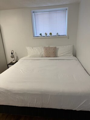 Ein ordentlich bezogenes Bett mit weißer Bettwäsche, ein Nachttisch mit Lampe und ein Fenster mit Jalousien und Topfpflanzen.