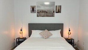 Ein ordentlich bezogenes Bett mit grauem Kopfteil, zwei Nachttische mit Lampen und ein Stadtbild über dem Bett.