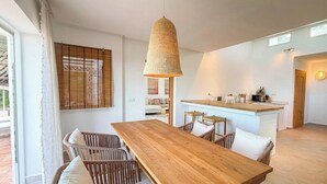 Een moderne keuken met een houten eiland, een eethoek met een grote houten tafel en rieten stoelen, en een hangende lamp.
