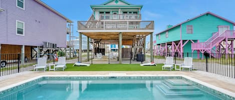 A swimming pool with a wooden deck, surrounded by a fence, and adjacent to a building with a balcony.