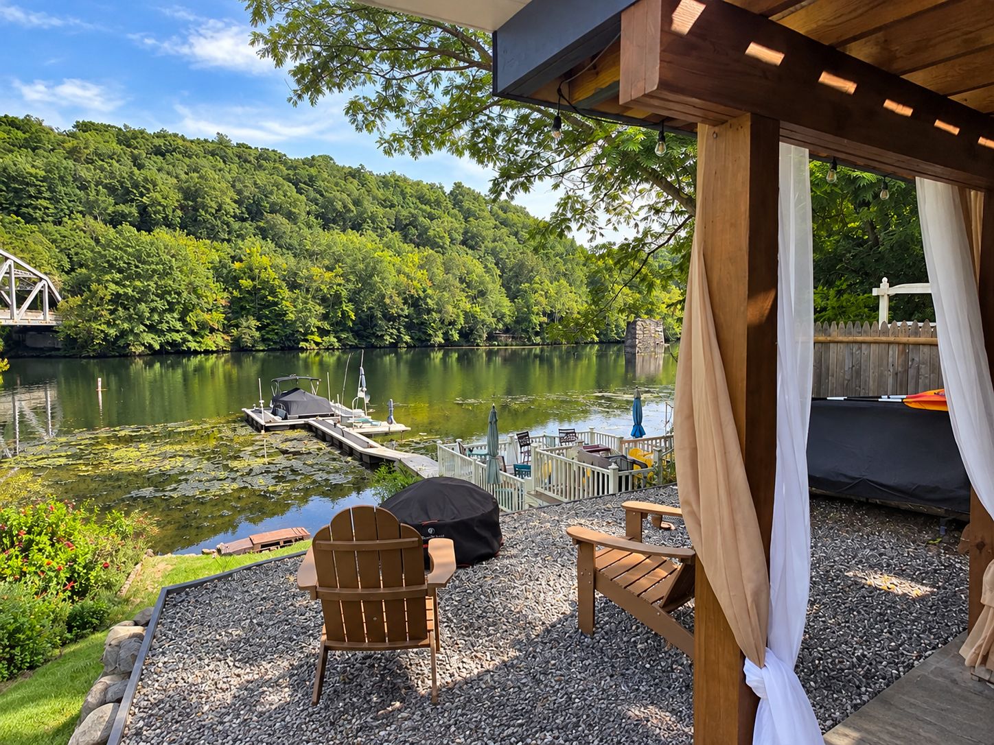 A lakeside patio with a wooden deck, a covered seating area, and a view of a forested hillside.