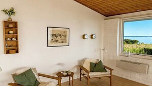 Ein Zimmer mit Holzboden, zwei Stühle mit grünen Kissen, ein kleiner Tisch, ein Korbwäsche und ein gerahmtes Bild, sowie ein Fenster mit Meerblick.
