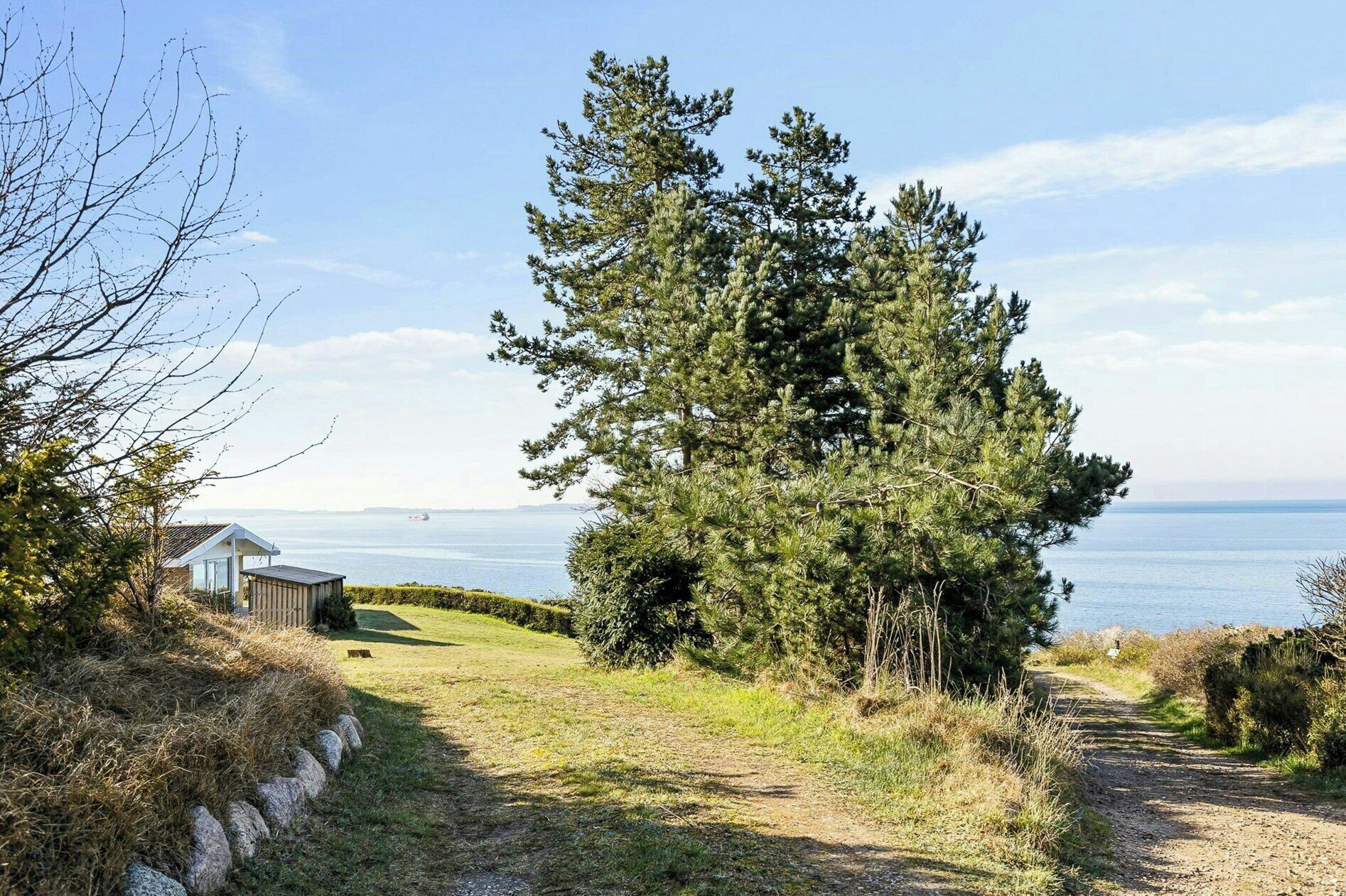 A coastal path with a small wooden structure, dense trees, and a clear view of the sea.