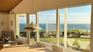 Ein stilvoller Essbereich mit Holztisch und weißen Stühlen, große Fenster mit Blick auf das Meer und eine Holzzielung.