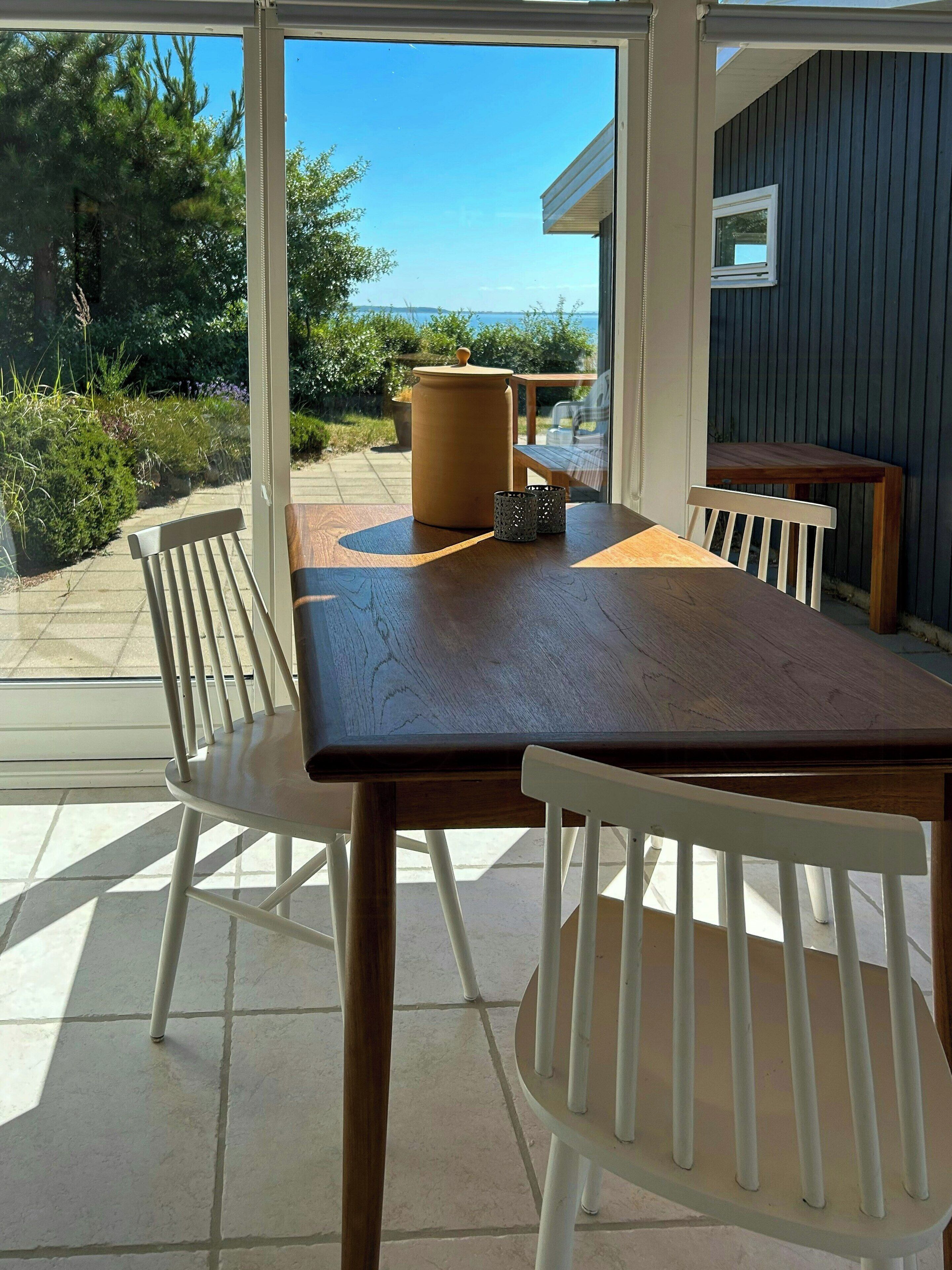 Une terrasse extérieure avec une table en bois et des chaises blanches, un grand pot de fleurs et une petite table placée près d’une fenêtre.