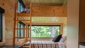 Interior de una cabaña con literas, una cocina con taburete y ventana con vistas a la vegetación.