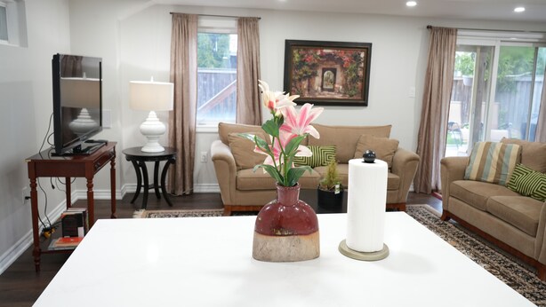 Ein Wohnzimmer mit einem weißen Couchtisch, einer roten Vase mit Blumen, einem weißen Teelicht, einem Fernseher auf einem Schrank und einem beigen Sofa mit einem grünen Kissen.