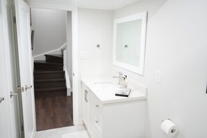 Une salle de bain avec un lavabo blanc, un miroir et un escalier en bois sombre menant à une autre pièce.