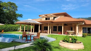 Una casa de dos pisos con piscina, mobiliario exterior y un patio cubierto.