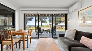 A modern living room with a wooden dining table, chairs, a dark sofa, and a view of the outdoors through large glass doors. - St. Leonards Bay Views (St Leonards)