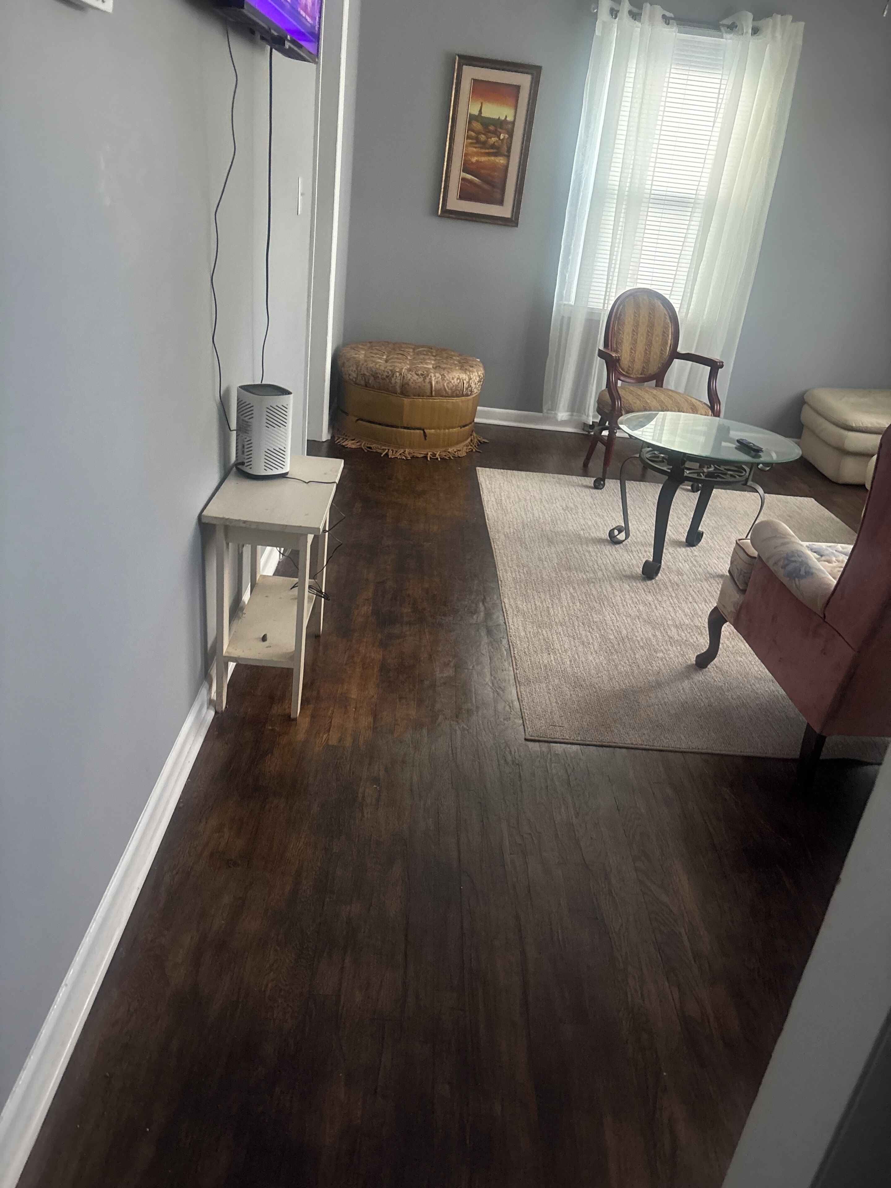 Un salon doté d’un parquet en bois foncé, d’une petite table blanche, d’un pouf rembourré, d’une table basse en verre, d’une chaise et d’un canapé.