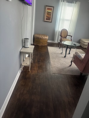 Una sala de estar con piso de madera oscura, una pequeña mesa blanca, un otoman acolchado, una mesa de centro de vidrio, una silla y un sofá.