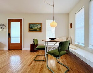 A dining area with a round table, two green chairs, a hanging light fixture, a painting on the wall, and a window with blinds. - Serene, charming 2-bed bungalow, filled with art and light, in Central Austin (Austin)