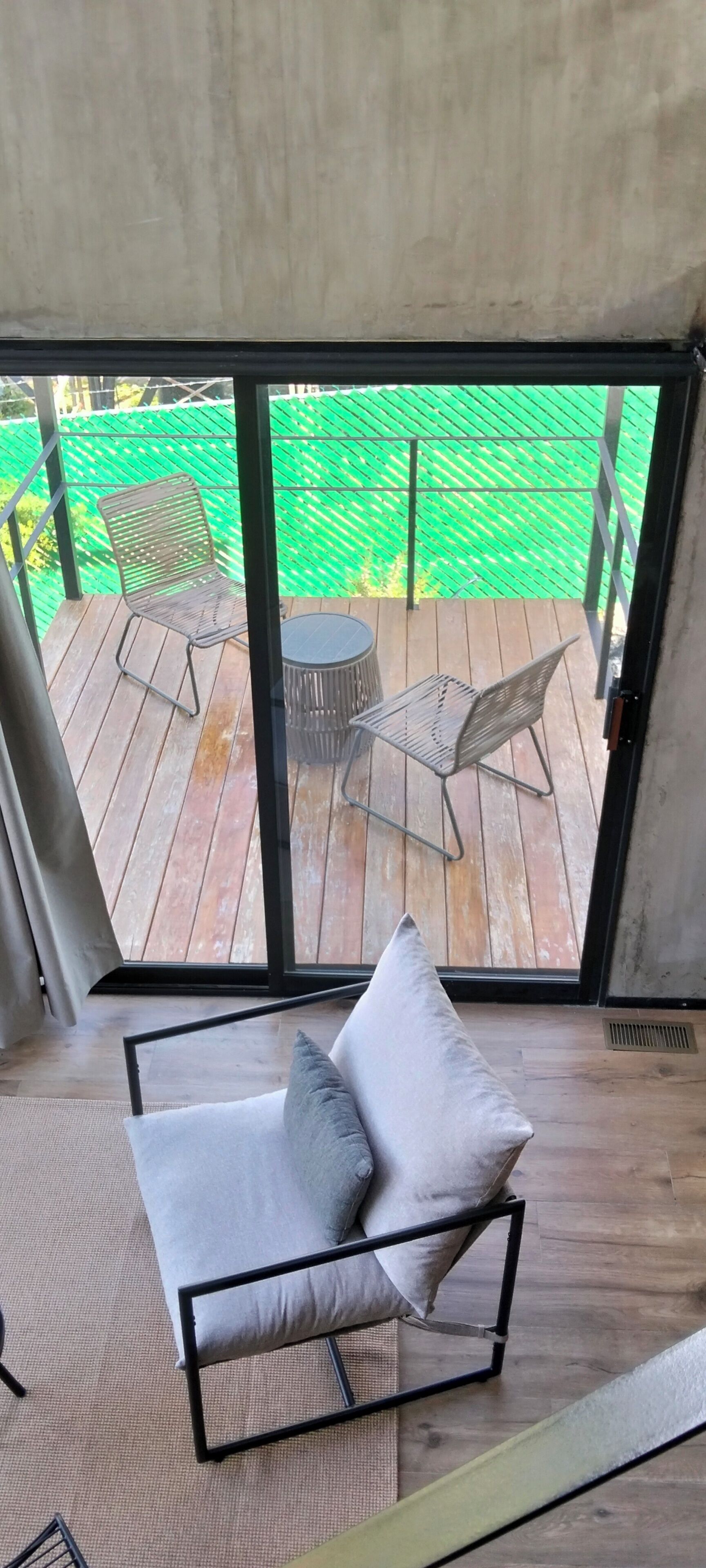 Een kamer met een bank, een klein tafeltje en uitzicht op een houten terras en een groene omheining.