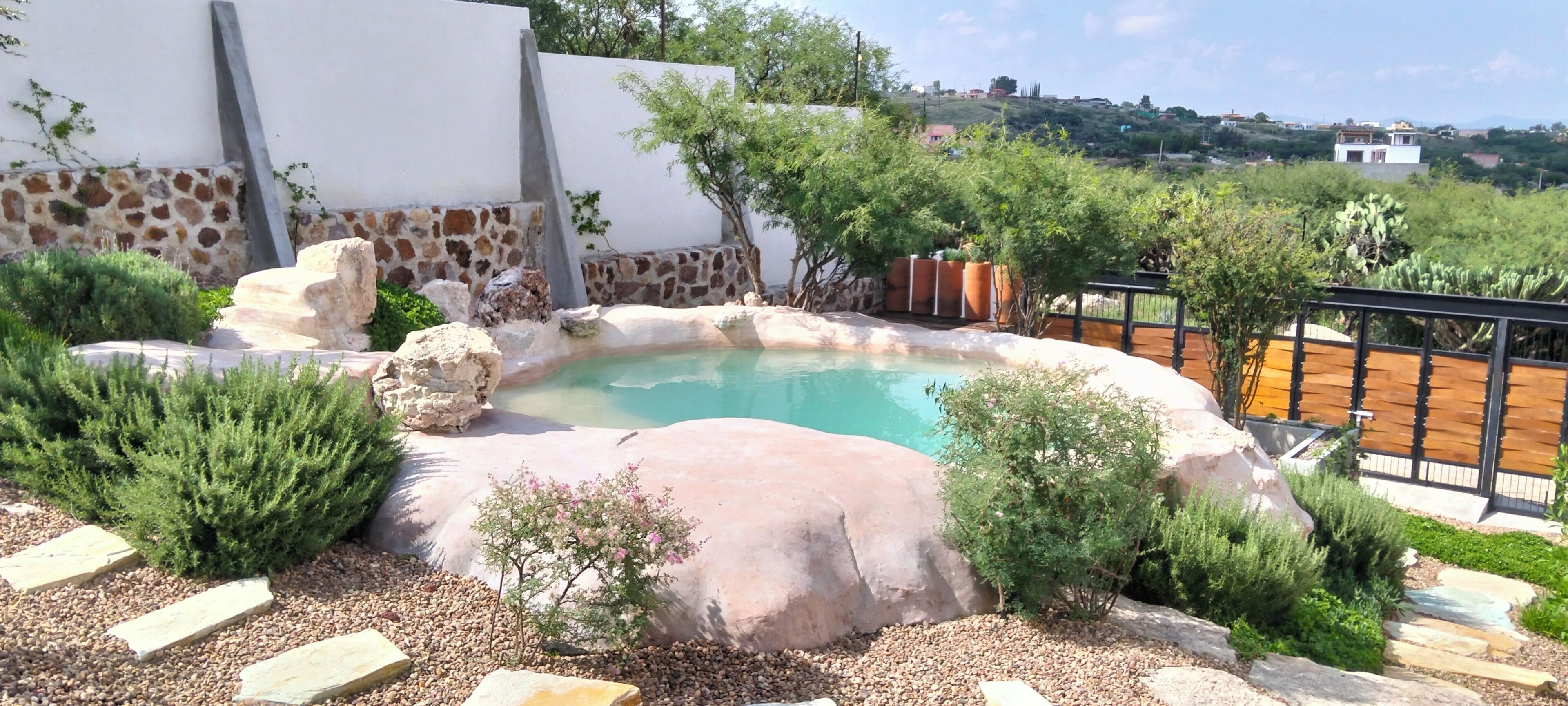 A pool surrounded by rocks and plants, with a white wall and a view of houses in the background.