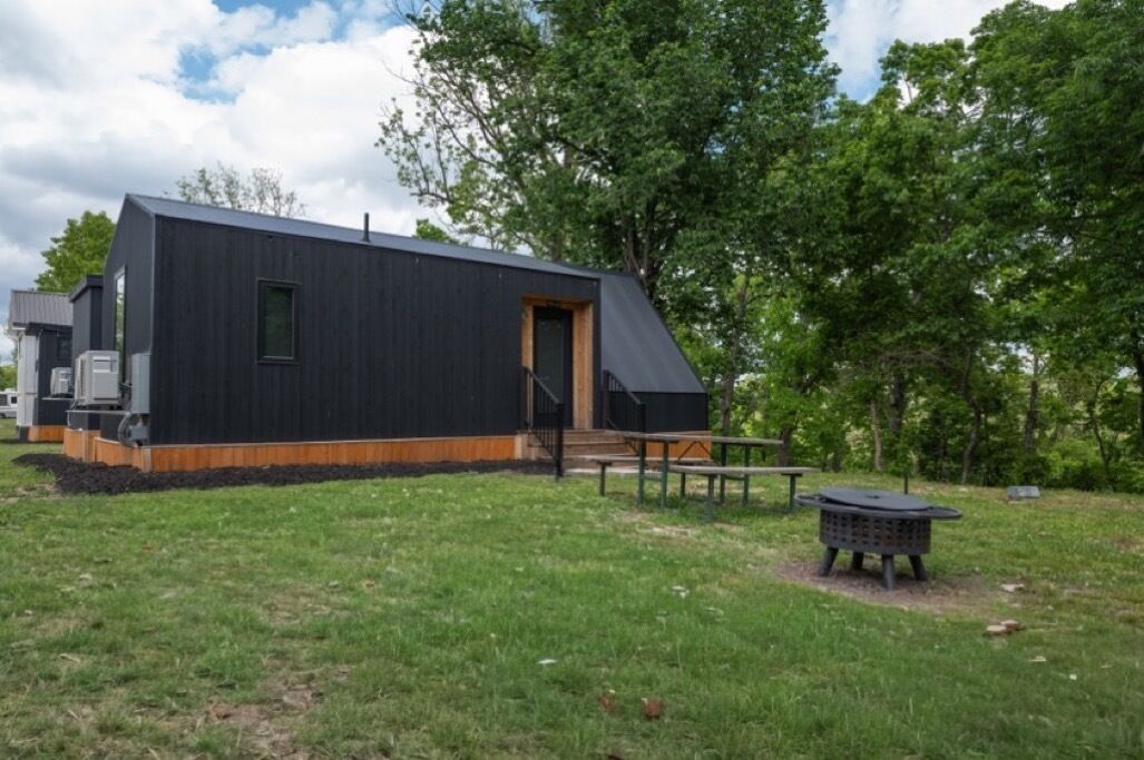 Ein modernes, schwarzes Gebäude mit geneigtem Dach, einer Holzterrasse und einem Picknicktisch in einer grünen Wiese.
