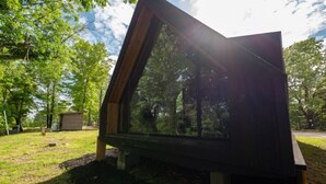 A modern, dark-colored structure with a unique triangular design, situated in a grassy area with trees in the background. - A-Frame Retreat at Beaver Lake - Sleeps 4 near Rogers & Bentonville (Rogers)