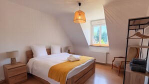 Un dormitorio con cama, mesitas de noche, lámpara colgante, ventana y una silla.