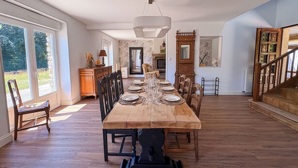 Un comedor amplio con una larga mesa de madera dispuesta para una comida, flanqueada por sillas, y una escalera que lleva a un nivel superior.