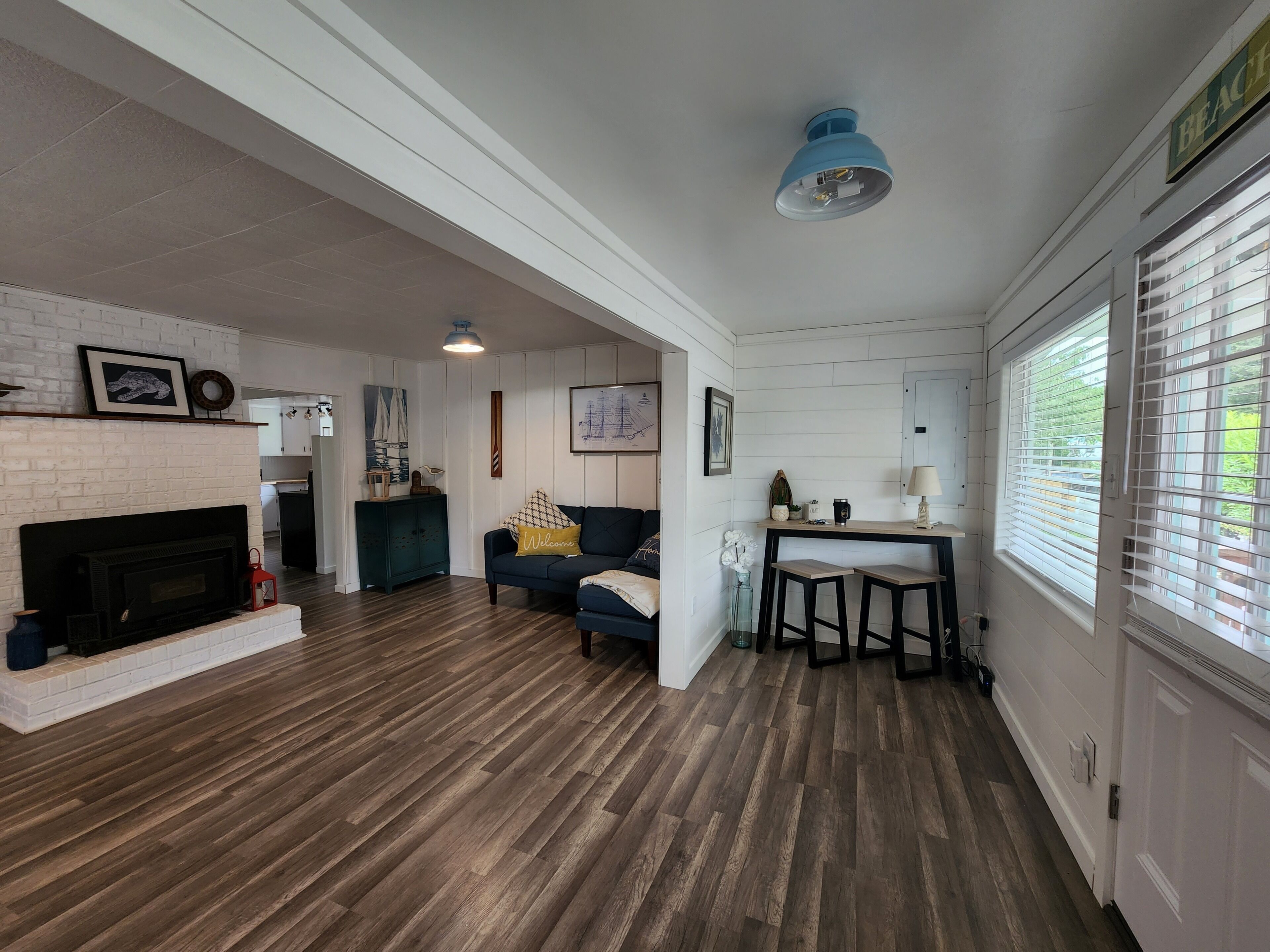 A spacious living area with a fireplace, wooden flooring, a blue ceiling light, and a kitchen area with a window.