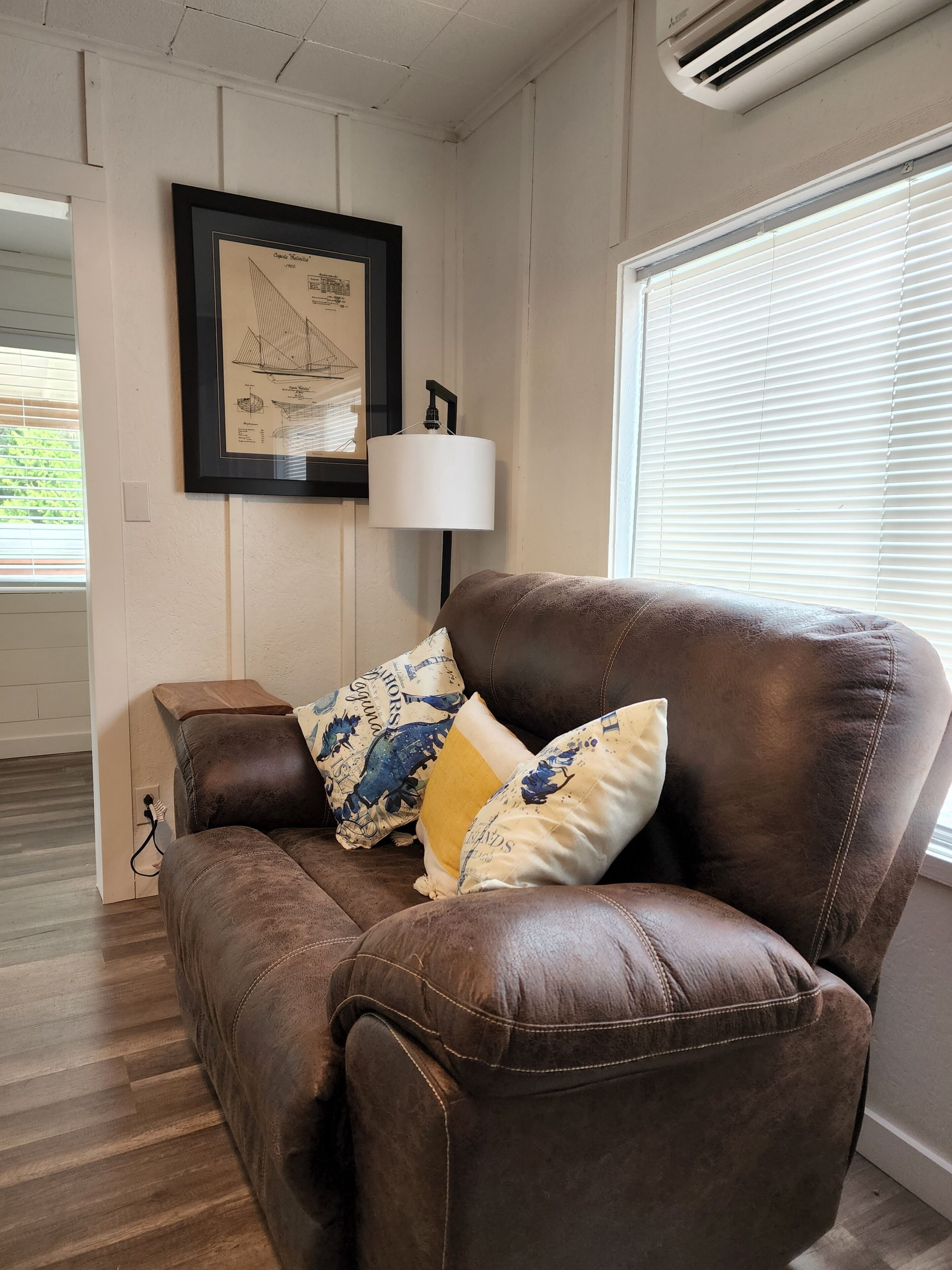 A brown leather sofa with patterned cushions, a framed architectural drawing on the wall, a floor lamp, and a window with blinds.