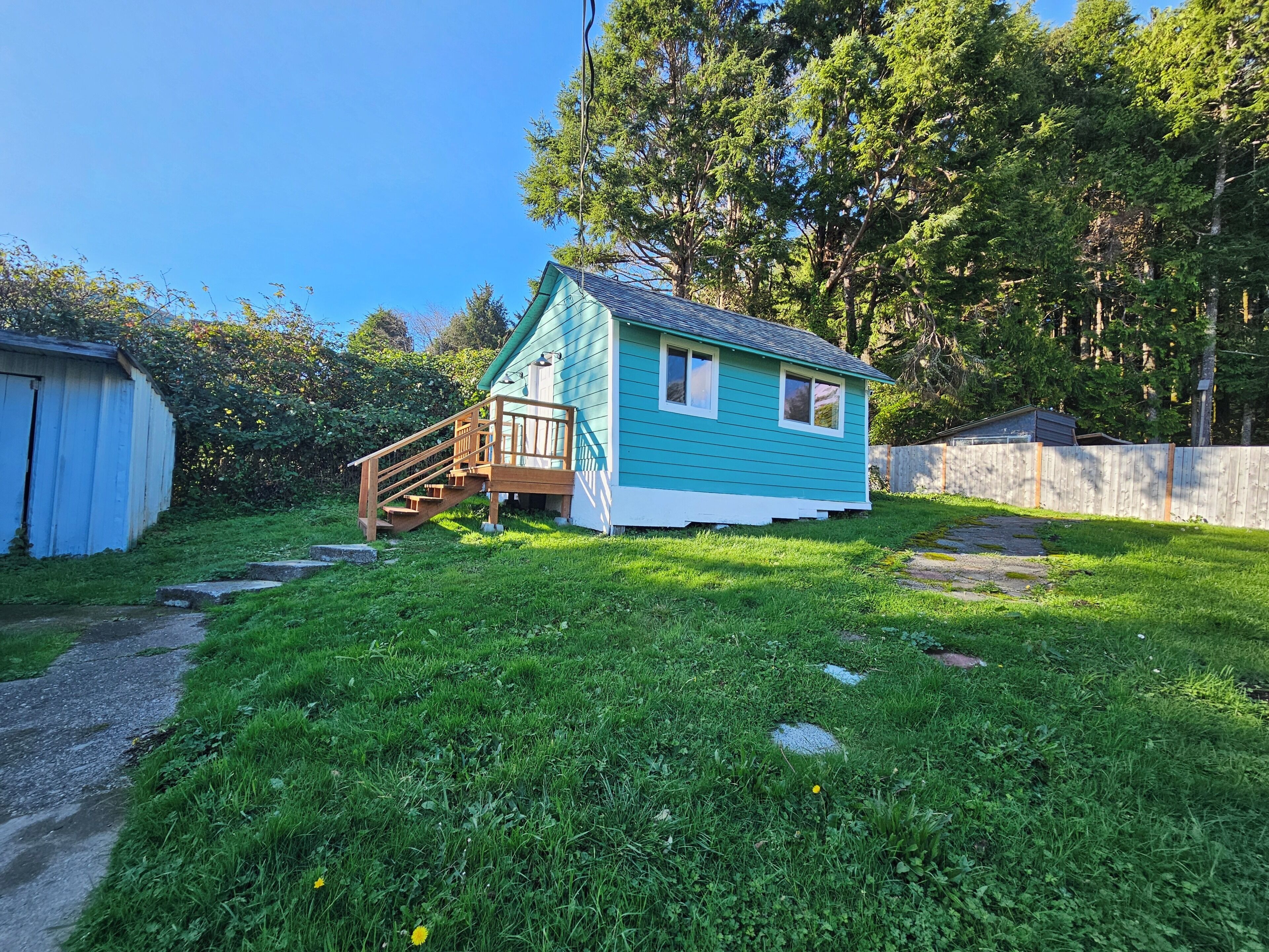 A small house with a wooden staircase leading to a porch, surrounded by a grassy yard and trees.