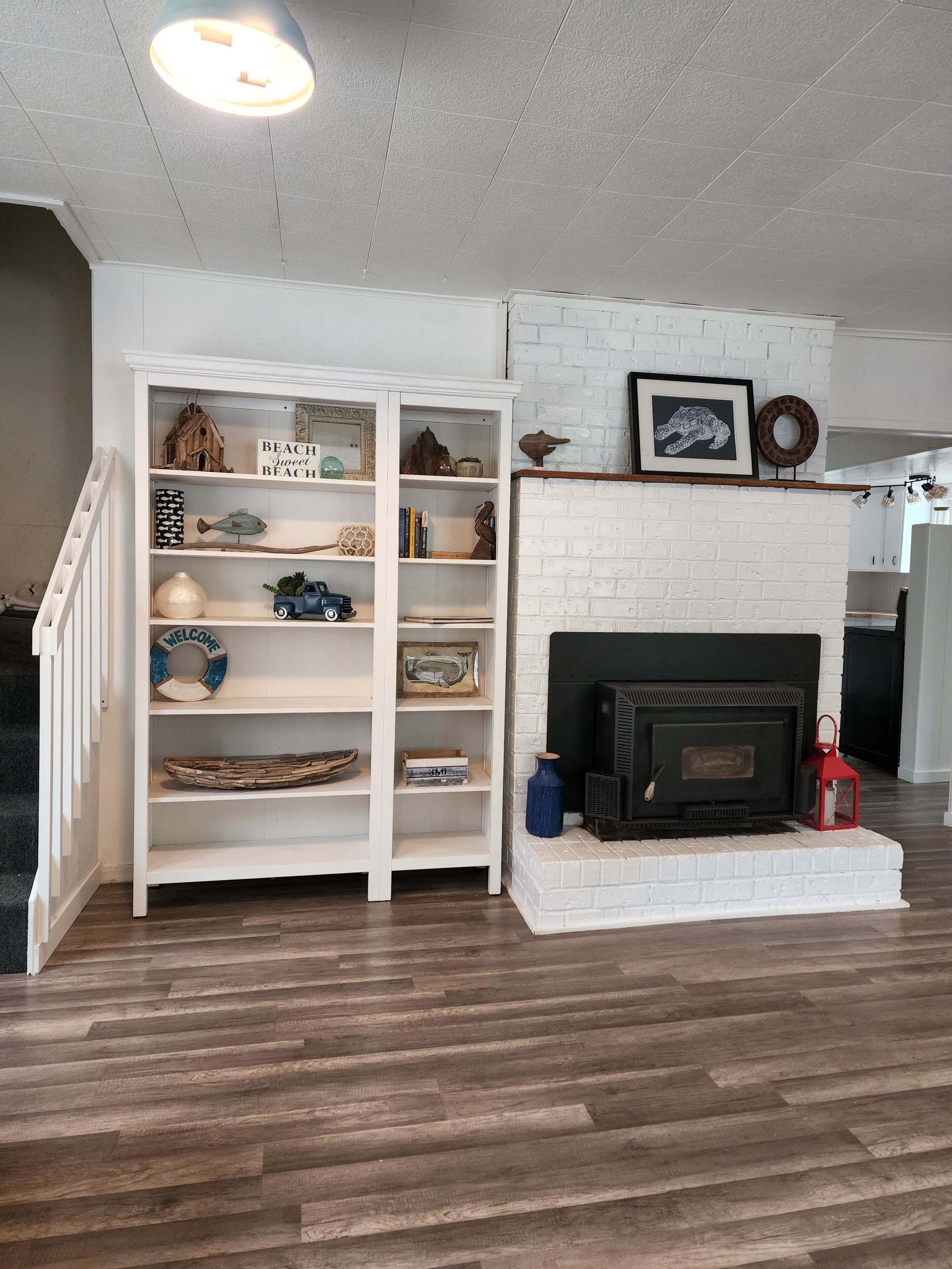 A living room with a white brick fireplace, a white bookshelf with various decorative items, and a staircase leading to another level.