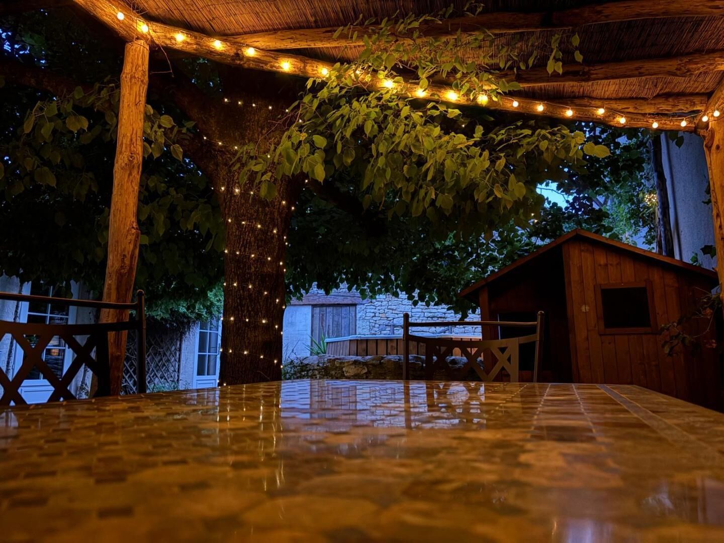 Un espace extérieur abrité, aménagé avec une table, des guirlandes lumineuses et une structure en bois.
