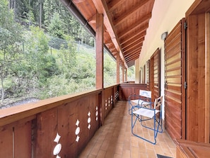 A wooden balcony with a view of a forested hillside, a table with chairs, and a door with a window. - Apartment Anny by Interhome (Canazei)