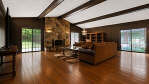 A spacious living room with a stone fireplace, wooden beams, and large windows.