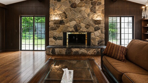 A spacious living room with a stone fireplace, wooden flooring, and large windows.