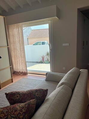 Ein Wohnzimmer mit einer Couch, gemusterten Kissen, einer Schiebetür zu einer Terrasse und Blick auf ein Gebäude.
