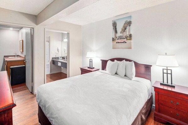 Una habitación de hotel con una cama, mesitas de noche, un escritorio, una silla, un espejo, un lavamanos y un cuadro de palmeras.