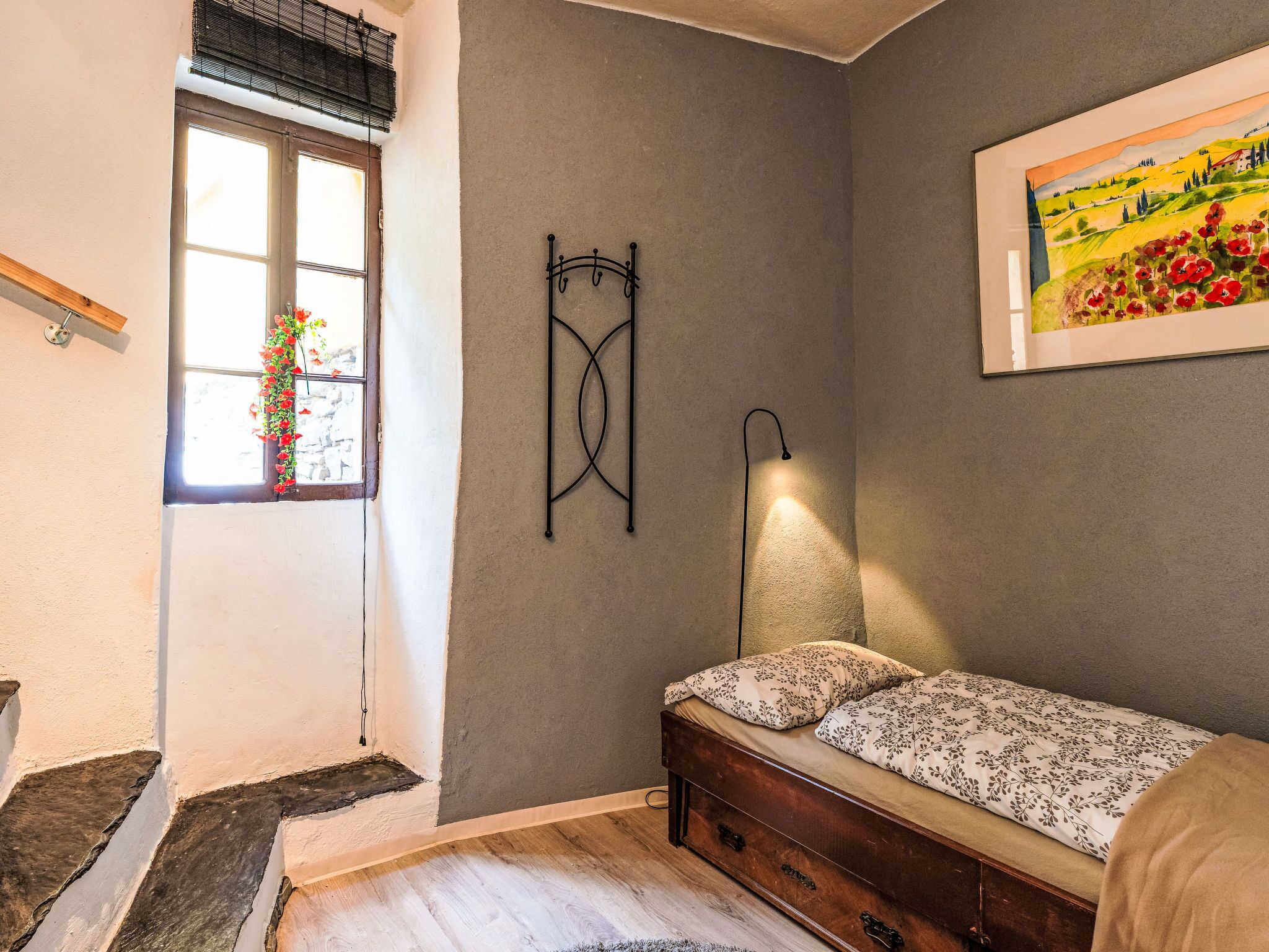 A bedroom with a bed, a nightstand, a wall-mounted lamp, a window with a view, and a painting on the wall.