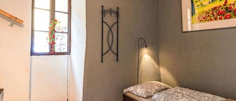 A bedroom with a bed, a nightstand, a wall-mounted lamp, a window with a view, and a painting on the wall.