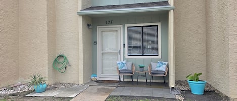 A building entrance with a door marked '177', a window with blinds, two wicker chairs with cushions, and two potted plants.
