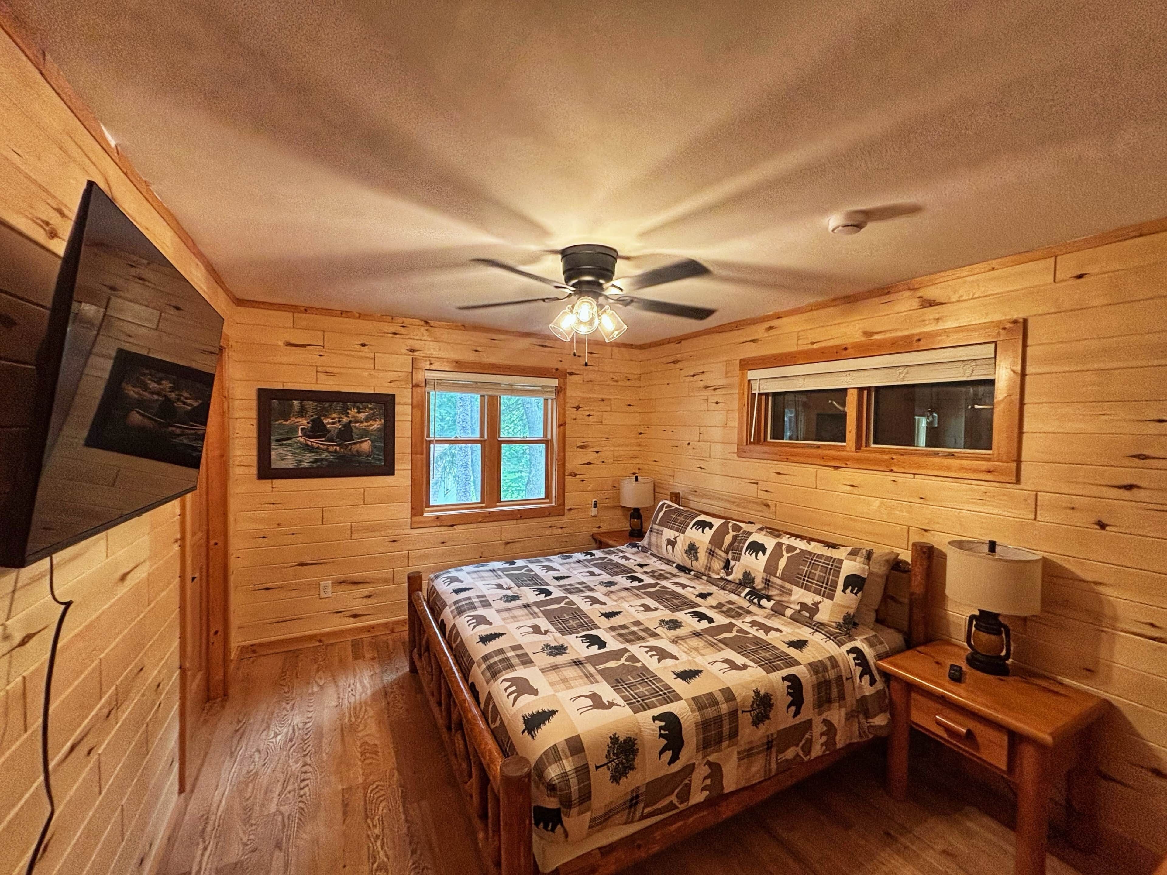 A wooden cabin bedroom with a bed, two windows, a ceiling fan, and a nightstand.
