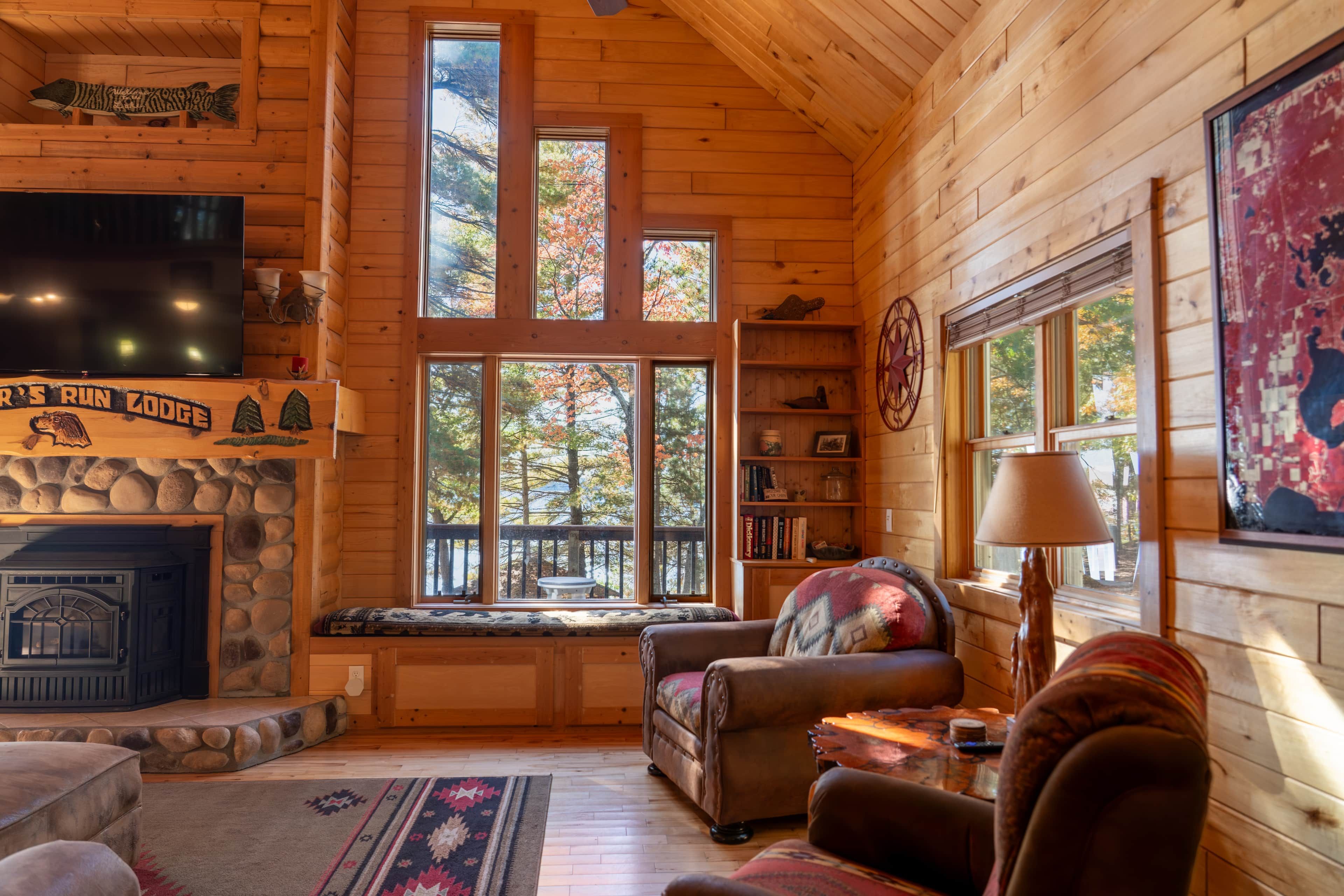 A cozy cabin interior with a stone fireplace, a television, a bookshelf, and a large window overlooking trees.