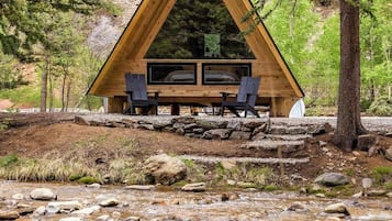 A wooden cabin with a triangular roof, two windows, and two chairs outside.
